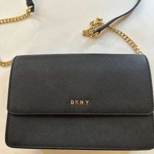 Svart axelväska från DKNY - Snygg svart axelväska från DKNY med guldfärgad kedja och justerbar axelrem i skinn. Väskan har stilren design med DKNY-logga i guld på framsidan och flera fack inuti. Perfekt storlek för det viktigaste och kommer med original dustbag. Använd ett fåtal gånger. Inga tecken på användning, repor eller rost. 