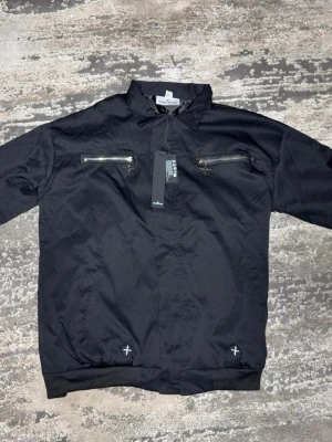 Svart bomberjacka från Stone Island - Svart bomberjacka från Stone Island med flera silvriga dragkedjor på bröstet, ärmen och ryggen. Jackan har klassisk krage, ribbade muddar och detaljer med vita korsstygn vid nederkanten. Materialet är polyester och jackan har en loose passform.