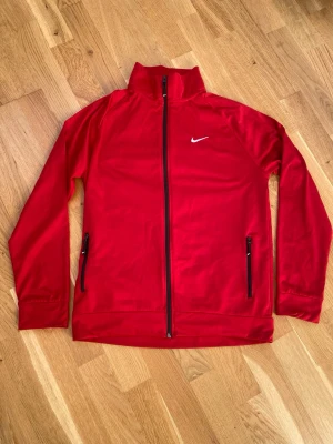 Röd Nike zip-tröja med hög krage - Snygg röd zip-tröja från Nike med hög krage och svart dragkedja hela vägen. Tröjan har två fickor med dragkedja och en vit Nike-logga på bröstet. Perfekt för träning eller chill. Materialet känns mjukt och lätt, och passformen är normal.