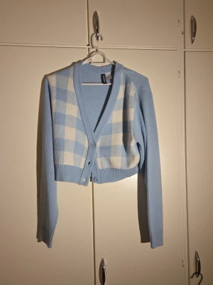 Blå rutig kofta från H&M Divided - Säljer en ljusblå croppad kofta från H&M Divided i storlek S. Koftan har ett rutigt mönster i vitt och blått framtill, långa ärmar och v-ringad hals med knappar. Materialet är mjuk och syntetisk akryl, perfekt för lager på lager under höst och vår.