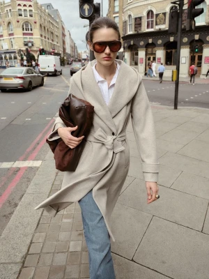 Beige kappa med huva och knytband - Stilren beige kappa med stor huva och knytband i midjan. Kappan har en avslappnad passform, långa ärmar och en enkel knapp framtill. Perfekt för dig som gillar en clean look med extra detaljer som markerad midja och bred krage.