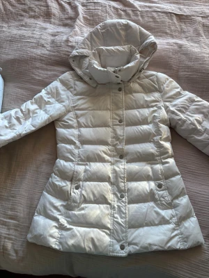 Vit pufferjacka från Tommy Hilfiger - Säljer en snygg vit pufferjacka från Tommy Hilfiger i storlek S. Jackan har huva, broderad logga på ärmen och knappar framtill. Den är quiltad och har en hög krage som skyddar mot kyla. Perfekt för dig som vill ha en stilren vinterlook.