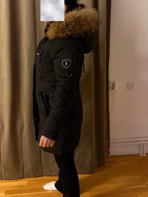 Svart lång dunjacka med päls från Rockandblue - Säljer en svart dunjacka från Rockandblue med lång passform och stor huva med fluffig brun päls. Jackan har flera dragkedjor, tryckknappar och snygga detaljer på ärmarna. Perfekt för kalla dagar med sin quiltade design och rejäla värme. Ett litet hål finns nere på fickan, inget som man lägger märke till så mycket! 