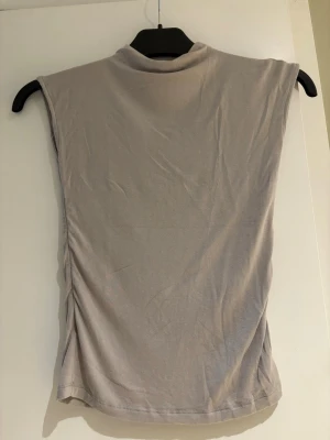 Beige ärmlös topp från Missy - Säljer en stilren beige topp från Missäy med hög hals och ärmlös design. Toppen har en mjuk och tunn känsla, perfekt för varmare dagar. Passformen är normal och materialet känns som bomull eller syntet. Enkel att matcha med jeans eller kjol.