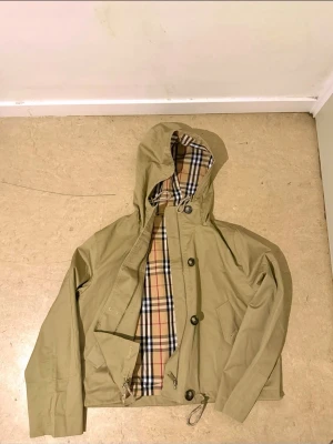 Burberry kort trenchcoat i beige - Burberrys korta parkajacka i beige med klassiskt rutigt foder, huva och snörning i midjan. Jackan har knappar och dragkedja framtill samt två fickor med knappstängning. Perfekt för dig som vill ha en clean och stilren look med ikonisk Burberry-vibe.