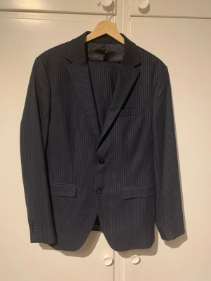 Mörkblå randig kostym  - Snygg mörkblå kostym från Riley med tunna vita ränder. Mycket bra skick,använd vid fåtal tillfällen.Perfekt för dig som vill ha en stilren och tidlös look. OBS!Kavaj-storlek 48,byxor 46.Går att säljas separat.Mycket finare i verkligheten än på bilden.