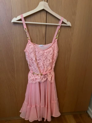 Rosa spetsklänning från Ida Sjöstedt - Supersöt rosa kortklänning från Ida Sjöstedt med spets upptill och volangig kjol. Klänningen har smala axelband med guldfärgade detaljer och ett matchande bälte i midjan. Materialet är lätt och luftigt, perfekt för varma dagar. Klänning är endast använd en gång. Strl står ej med på lapp, men passar en S.