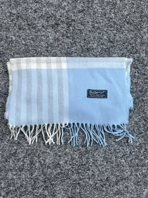 Ljusblå rutig halsduk från Burberry - Schysst ljusblå halsduk från Burberrys med klassiskt rutigt mönster i blått, vitt och grått. Halsduken har fransar i ändarna och en diskret Burberrys-label. Tillverkad i 100% lammull, mjuk och skön att ha runt halsen.