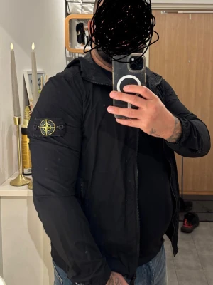 Svart Stone Island vindjacka med huva - Svart vindjacka från Stone Island med klassisk patch på vänster ärm. Jackan har huva, dragkedja framtill och flera fickor på insidan. Tillverkad i nylon som ger en lätt och skön känsla. Perfekt för dig som gillar streetwear och vill ha en clean look.passar bra till vardags och finare tillfällen, jacka passar inte mig längre. 