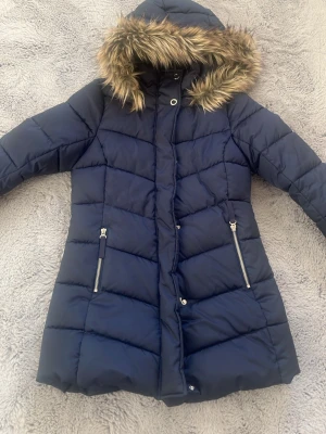 Mörkblå pufferjacka med päls från H&M - Snygg mörkblå pufferjacka från H&M med huva och päls. Jackan har långa ärmar, två fickor med dragkedja och quiltad design. Perfekt för kalla dagar och har en skön, varm känsla. Stängs med både dragkedja och knappar framtill.