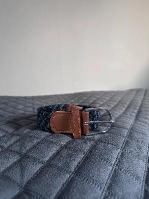 Svart grått blått flätat bälte - Tja det är helt nytt flätat bälte från Bältgallerian. Det är 105cm långt och 3,5cm brett. Det är i ett elastiskt material så det passar mindre storlekar också 
