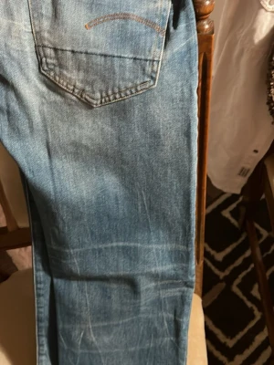 Blå jeans från G-Star RAW - Säljer ett par klassiska blå jeans från G-Star RAW med snygga slitningar och kontrastsömmar. Jeansen har raka bakfickor med broderade detaljer och en stor läderpatch i midjan. Perfekta till sneakers och hoodie för en chill look.