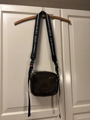 Svart axelväska från M+RC Noir - Säljer en svart axelväska från M+RC Noir med transparent framsida och präglad logga över hela väskan. Väskan har bred, justerbar rem med vita detaljer och metallspännen i regnbågsfärgad finish. Perfekt för dig som gillar streetwear och vill sticka ut.