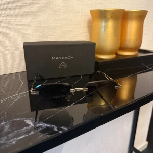 Maybach solglasögon med silverdetaljer - Exklusiva Maybach solglasögon med kantig form och tunna silverfärgade detaljer. Glasen är mörkgrå och bågarna har en lyxig design med Maybach-logga på sidorna. Handgjorda i Tyskland, perfekt för dig som gillar stil och kvalitet.