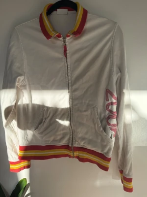 Vit zipperhoodie Porsche Design - Snygg vit bomberjacka från Porsche Design med '2006' på ärmen och stort Porsche-tryck på ryggen samt armen. Jackan har dragkedja, ribbade muddar och krage i rött och gult, samt två fickor fram. Materialet är mjuk bomull och trogen till sin storlek :)