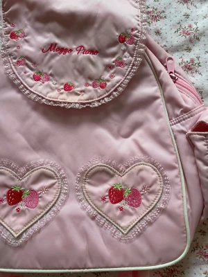 Rosa ryggsäck från Mezzo Piano med jordgubbar - Supergullig rosa ryggsäck från Mezzo Piano med broderade jordgubbar och hjärtan på framsidan. Ryggsäcken har spetsdetaljer, justerbara axelband och dragkedja i matchande rosa. Köpt i Japan och aldrig använd! 