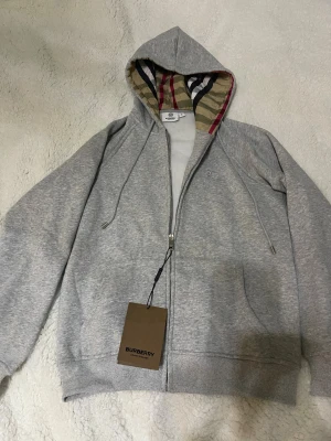 Grå hoodie från Burberry med dragkedja - Snygg grå hoodie från Burberry med klassiskt rutigt foder i huvan och broderad logga på bröstet. Tröjan har dragkedja, snörning i huvan och ribbade muddar. Perfekt för dig som gillar stilrena detaljer och exklusiva märken.