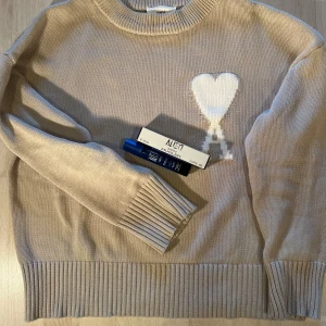 Kakhi stickad tröja från Ami Paris - En parfym på köpet! Snygg beige stickad tröja från Ami Paris med ett vitt hjärta och bokstaven A broderat på bröstet. Tröjan har rund hals, långa ärmar och breda ribbade muddar vid ärmslut och nederkant. Perfekt för dig som gillar stilrena detaljer och mjuka material. 