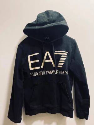 Emporio Armani - Svart hoodie från EA7 Emporio Armani med stor vit logga framtill och mindre logga baktill. Tröjan har huva med snörning, känguruficka och långa ärmar. Tillverkad i mjuk bomull, perfekt för chill dagar och streetwear-stil.