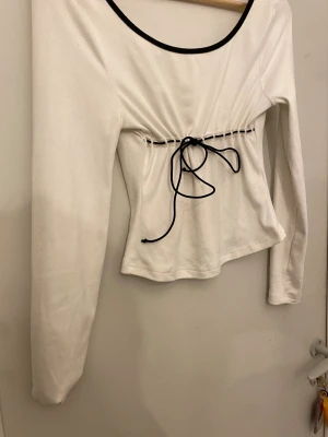 Vit långärmad topp med snörning - Säljer en vit långärmad topp med svart kant runt halsen och snygg snörning i midjan. Toppen har en rundad halsringning och är figurnära med en lätt croppad passform. Perfekt för dig som gillar kontraster och stilrena detaljer.
