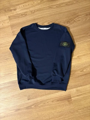 Marinblå Stone Island sweatshirt - Snygg marinblå sweatshirt från Stone Island med klassisk rund hals och ribbade muddar. Ikonisk patch med logga på vänster ärm, fästs med två svarta knappar. Tröjan är gjord i mjuk bomull och har en clean, normal passform som funkar till jeans eller cargo.