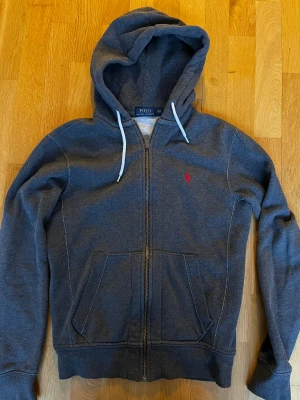 Mörkgrå hoodie från Polo Ralph Lauren - Snygg mörkgrå hoodie från Polo Ralph Lauren med vit snörning i huvan och röd broderad logga på bröstet. Tröjan har dragkedja framtill, två fickor och är tillverkad i mjuk bomullsmix. Perfekt för chill dagar och streetstyle.