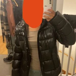 Svart pufferjacka med huva - Säljer en svart pufferjacka med glansig finish och huva. Jackan har långa ärmar, dragkedja och knappar framtill. Perfekt för kalla dagar och har en oversized look som är riktigt trendig just nu. Hål på som är lagat hos sömmerska 
