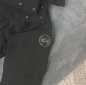 Svart dunjacka från Canada Goose - Svart dunjacka från Canada Goose med huva och klassisk logga på ärmen. Jackan har dragkedja och knappar framtill, samt flera fickor. Materialet är slitstark polyester och dunfyllning som håller dig varm under kalla dagar. Perfekt för dig som vill ha en stilren och funktionell vinterjacka.