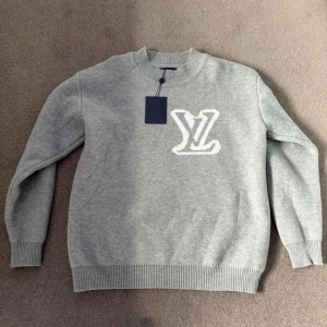 Grå stickad tröja Louis Vuitton - Snygg grå stickad tröja från Louis Vuitton med stort vitt LV-logo framtill. Tröjan har rund hals, ribbade muddar och långärmad design. Perfekt för dig som gillar exklusiva märken och stilren look.