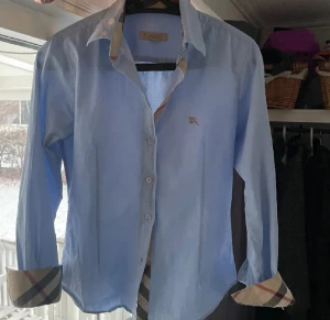 Blå Burberry Brit skjorta, S - Snygg ljusblå skjorta från Burberry Brit i storlek S. Dock är den lite liten för s så skulle mer säga xs eller xss.  Klassisk krage och långa ärmar med ikoniska rutiga detaljer på insidan av manschetter och krage. Skjortan har knappar framtill och ett litet Burberry-emblem broderat på bröstet. Perfekt för dig som gillar stilrena plagg med lyxig touch. 