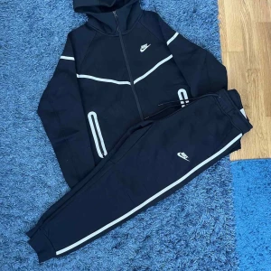 Svarta nike tech reflective - Snygga svart reflective tracksuit från Nike med vita ränder längs benen och logga på låret. Byxorna har snörning i midjan och ribbade muddar vid bensluten. Perfekta för chill eller träning, riktigt skön passform och stilren look.