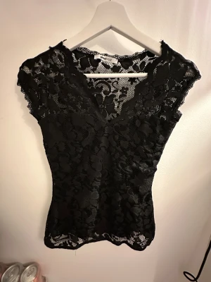 Svart spetsblus från Gina Tricot - Säljer en svart blus från Gina Tricot i storlek S. Toppen är helt i spets med blommigt mönster, v-ringad hals och ärmlös design. Den har en slim passform och transparenta detaljer som ger en snygg look. Perfekt för dig som gillar stilrena och trendiga plagg.