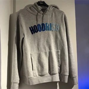 Grå hoodie från Hoodrich med blå text - Säljer en grå hoodie från Hoodrich i storlek M. Tröjan har en stor magficka, huva med snörning och broderad HOODRICH-text i olika blå nyanser på bröstet. Perfekt för chill dagar och streetwear-stil.
