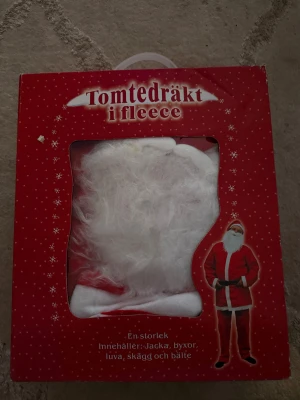Tomtedräkt i fleece med skägg - Tomtedräkt i mjuk fleece, komplett med röd jacka, byxor, vit luva, vitt skägg och svart bälte. Perfekt för att klä ut sig till tomte under julen. Dräkten har klassisk röd färg med vita detaljer och är bekväm att bära.