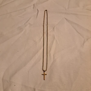 Guldigt halsband med kors - Snyggt halsband i guldton med en enkel korsberlock. Kedjan är klassisk och har ett smidigt lås. Perfekt för dig som gillar stilrena accessoarer med lite edge. Korset är slätt och minimalistiskt, vilket gör det lätt att matcha med andra smycken.