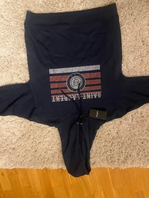 Marinblå hoodie från Saint Laurent - Snygg marinblå hoodie från Saint Laurent med tryck i rött, vitt och grått framtill. Tröjan har huva med snörning och en stor magficka. Materialet är mjuk bomull och passformen är normal. Perfekt för dig som gillar streetwear och exklusiva märken.