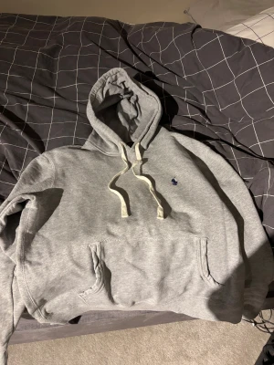 Grå hoodie från Polo Ralph Lauren - Snygg grå hoodie från Polo Ralph Lauren med klassisk blå logga broderad på bröstet. Tröjan har huva med vita snören, stor magficka och ribbade muddar vid ärmslut och nederkant. Perfekt för chill dagar och enkel att matcha med jeans eller joggers.