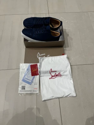 Mörkblå Christian Louboutin sneakers - Säljer ett par mörkblå sneakers från Christian Louboutin med ikoniska röda sulor och coola nitar på tån. Skorna har snörning och är tillverkade i mocka med vita platta sulor. Perfekt för dig som vill sticka ut med lyxig streetstyle. Dem är använda typ en hel skolvecka, pris kan diskuteras vid snabbaffär. Boxen är lite sliten skickar bild på boxen ifall du är intresserad av att köpa.