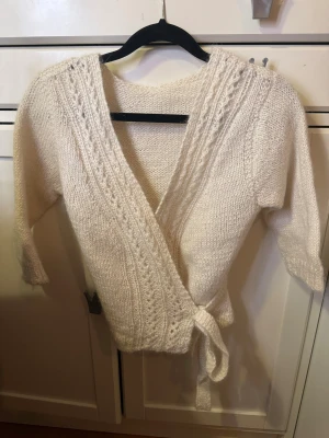 Vit omlottstickad kofta med knytband - Mysig vit stickad kofta med omlottmodell och knytband i midjan. Koftan har trekvartsärmar och ett snyggt ribbat mönster längs kanten.  Materialet är mjukt och fluffigt, koftan är hemstickad i strl XS-S