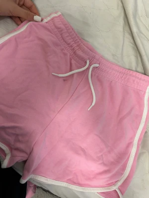 Rosa sportiga shorts med vita detaljer - Säljer ett par ljusrosa shorts från Cubus i storlek 158/164. De har vita snören i midjan och vita kantband längs sidorna. Modellen är loose och har elastisk midja med snörning, perfekt för chill eller träning. Materialet är mjuk bomull som känns skönt mot huden.