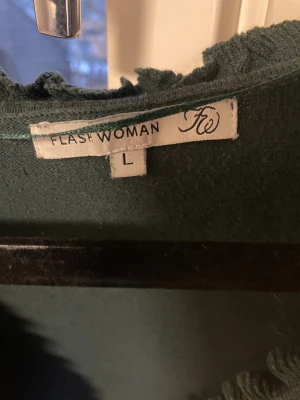 Mörkgrön kofta med volang från Flash Woman - Snygg mörkgrön kofta från Flash Woman i storlek L. Koftan har volangdetaljer längs v-ringningen och nederkanten, samt knappar framtill. Långärmad och tillverkad i mjukt stickat material som känns skönt mot huden. Det står storlek L men den passar mer en M.