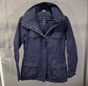 Jacka - Snygg marinblå vindjacka från Henri Lloyd med hög krage och flera praktiska fickor med dragkedja och lock. Jackan har lång ärm, justerbar midja och stilren design som funkar till många outfits. Perfekt för blåsiga dagar och enkel att matcha. Kan sänka pris vid snabb affär 