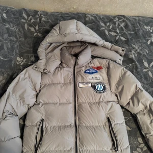 Jacka Mercier - Snygg grå pufferjacka från Mercier i storlek XS med huva och coola patchar på bröstet. Jackan har dragkedja framtill och två sidofickor med dragkedja. Perfekt för kalla dagar och har en modern, puffig look.