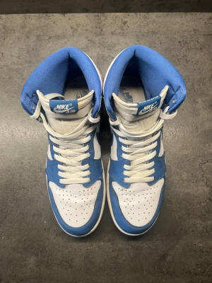 Nike Air Jordan 1 High OG University Blue - Nike Air Jordan 1 High OG i färgen University Blue. Skorna har en snygg kombination av vitt, ljusblått och grått skinn, klassisk snörning och den ikoniska swoosh-loggan på sidan. Perfekta för dig som vill sticka ut med en tidlös och sportig sneaker.