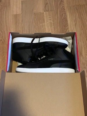 Svarta sneakers från Puma - Snygga svarta sneakers från Puma med vit platt sula och klassisk logga på sidan. Skorna har snörning och är tillverkade i en mix av skinn och syntetmaterial. Perfekt för dig som gillar stilrena och sportiga skor med en modern vibe. Det är skor anpassat till kallt väder som vinter. Dom är helt nya och har aldrig används förut