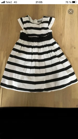 Randig vit och svart klänning med rosett - Snygg kortklänning från Kids by Lindex med breda svarta och vita ränder, rund hals och korta ärmar. Midjan har ett svart band med rosett och kjolen är lätt utställd med ett lager tyll under för extra volym. Dragkedja i ryggen för enkel påklädning.