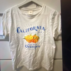 Vit California t-shirt från H&M - Snygg vit t-shirt från H&M med färgglatt tryck av apelsiner och texten 'California citrus fruit Oranges'. Klassisk rund hals och korta ärmar. Materialet är mjuk bomull som känns skönt mot huden. Säljer den eftersom jag inte använder den, har nästan aldrig haft på mig den så jag skulle säga att den är i ett ganska bra skick.