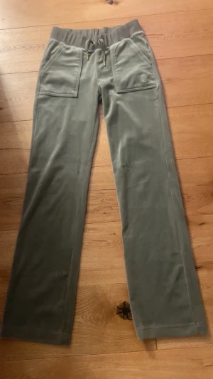 Juicy Couture XS - Snygga grönaktiga mjukisbyxor från Juicy Couture i storlek XS. Byxorna har raka ben, bred resår i midjan med snörning och guldfärgade detaljer. Fickor fram och bak med dekorativa knappar och broderad logga på ena fickan. Knappt använda, som nya.