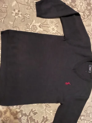 Polo Classic - Svart stickad tröja från Polo Classic med v-ringad hals och långärmad design. Tröjan har en röd broderad logga på bröstet och ribbade muddar vid ärmslut och nederkant. Perfekt för dig som gillar stilrena och klassiska plagg. Bara att det är i barn storlek men i manligt storlek är det mellan S och XS