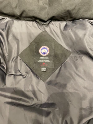 Svart dunjacka Canada Goose med päls - En exklusiv och klassiskt designad svart dunjacka från Canada Goose, skapad för att leverera maximal värme och komfort även under de kallaste vinterdagarna. Jackan är fylld med premiumdun som ger optimal isolering utan att kännas tung. Den har en robust ytterväv som står emot vind, kyla och lättare nederbörd.  Den ikoniska huvan är försedd med äkta päls som ger extra skydd mot snö och blåst, samtidigt som den ger jackan ett elegant och tidlöst utseende. Jackan är utrustad med praktiska fickor